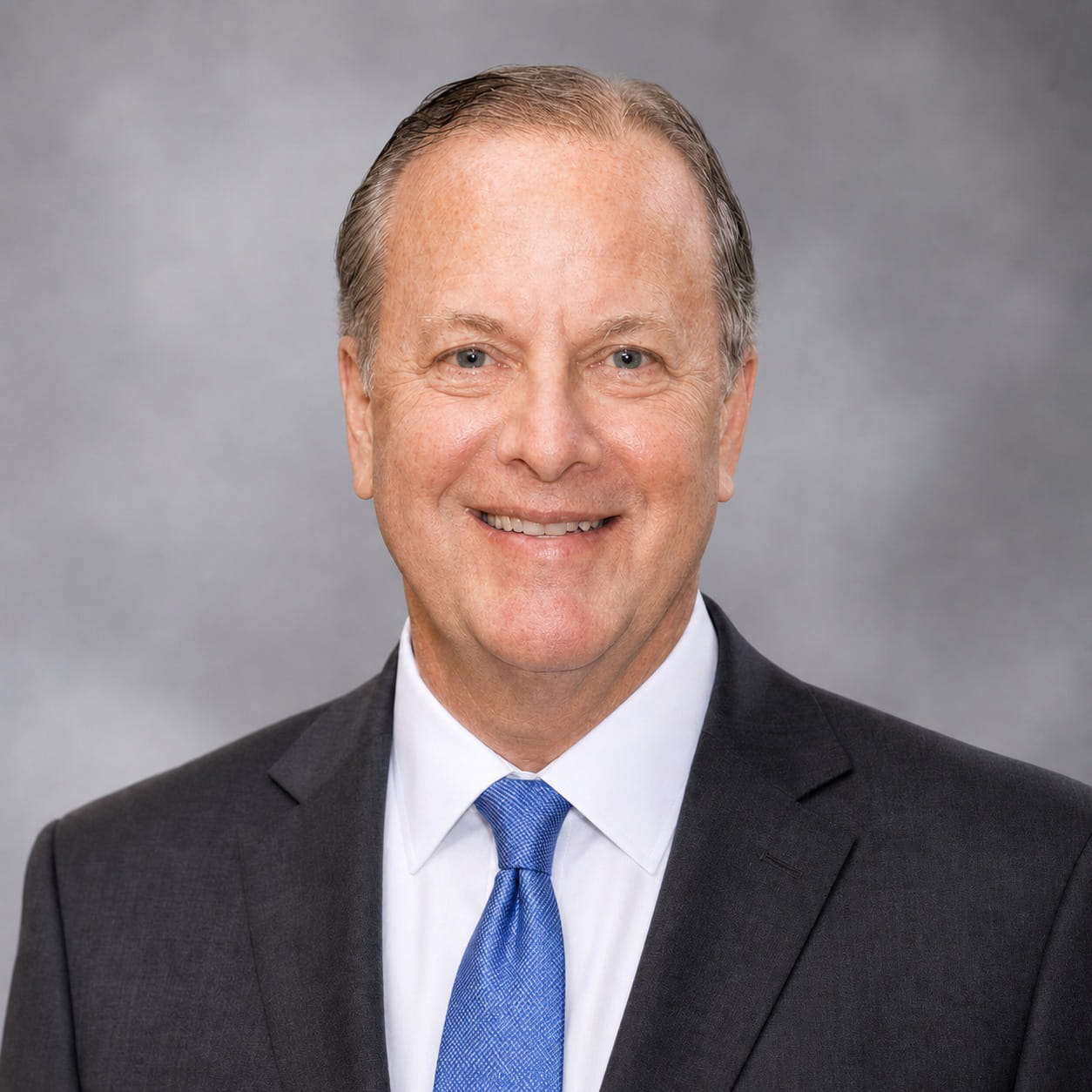 Douglas B. Baker wearing a dark suit, white shirt, and blue tie smiles in front of a neutral gray background.