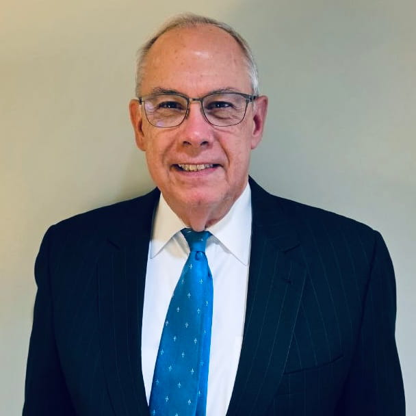 Ed Wilson, wearing a dark pinstripe suit, white shirt, and blue tie, stands against a plain light-colored background.