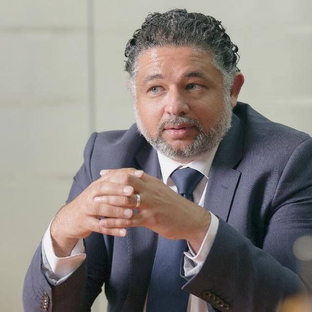 Mohammed Maghraby wearing a suit and tie, sits at a table with hands clasped, looking slightly to the side.