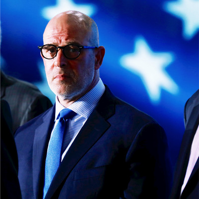 Ambassador Adam Ereli in a suit and glasses stands in front of a blurred background with large white stars on a blue backdrop.