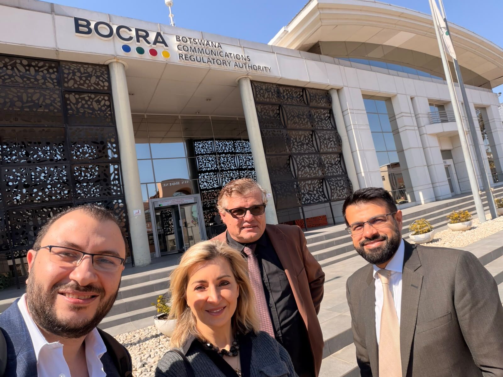 Four people in business attire stand smiling in front of the Botswana Communications Regulatory Authority (BOCRA) building on a sunny day.