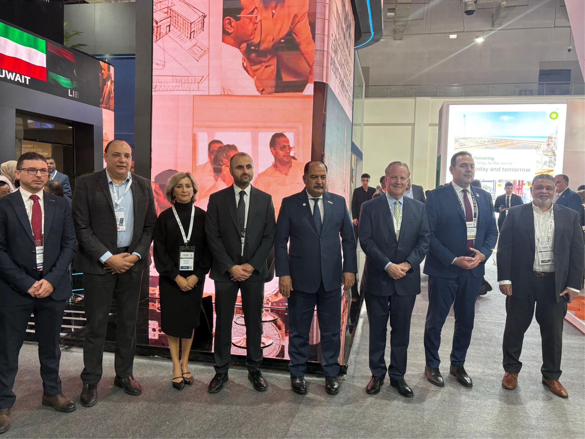 Eight people in formal business attire stand side by side at an indoor exhibition or conference, with display booths and screens in the background showcasing a variety of professional services.