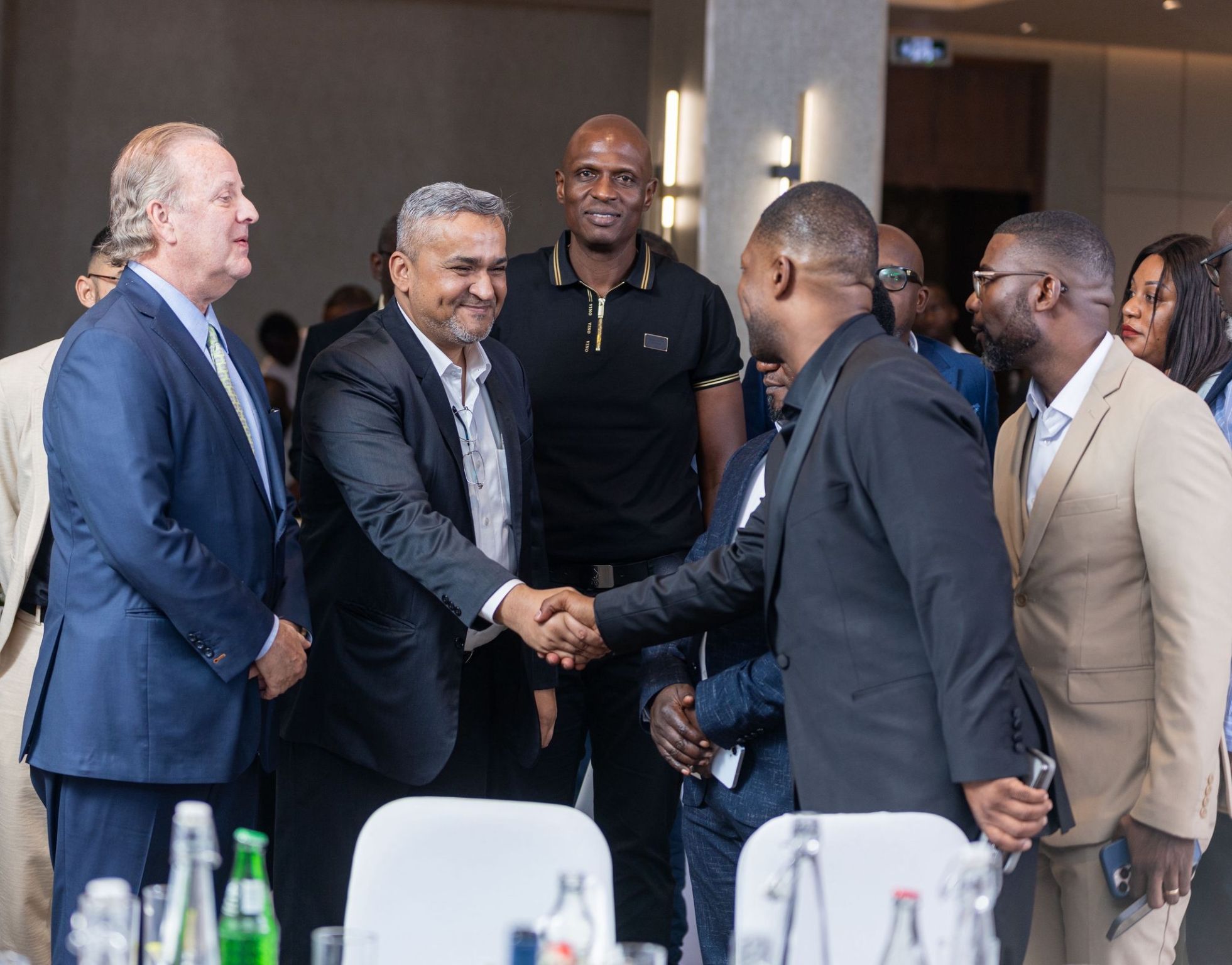 A group of people in business attire are gathered indoors, with two men in the center shaking hands across a table.
