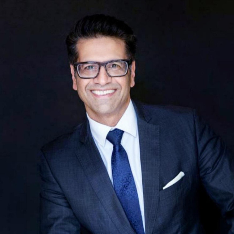 Ejaz Adham in a dark suit, white shirt, and blue tie smiles at the camera against a plain dark background.