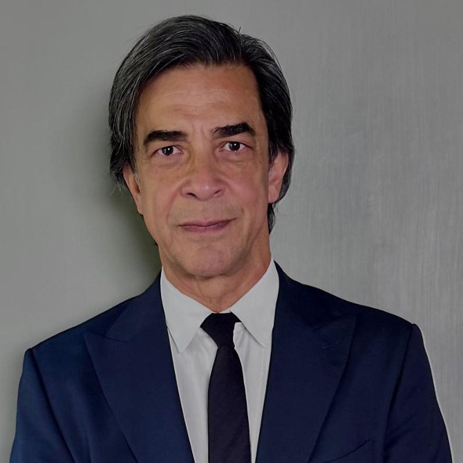 Guven Giray wearing a dark suit, white shirt, and black tie stands against a plain gray background.