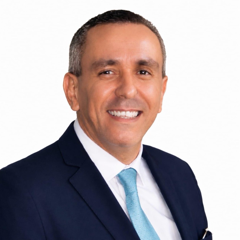 Hassan Abassi in a navy suit, white shirt, and light blue tie, facing forward against a plain white background.
