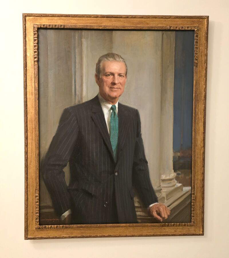 Framed oil painting of U.S. Secretary of State James A. Baker III in a dark pinstripe suit, white shirt, and teal tie, standing indoors with one hand resting on a table.