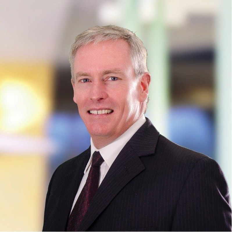 Paul Smith in a dark suit and tie is smiling at the camera, standing in front of a blurred indoor background with yellow and blue tones.